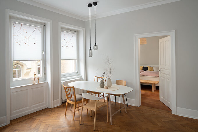 Zwei weisse Fenster mit floral bedruckten Wabenplissees in einem hellen Esszimmer mit Holzfussboden.
