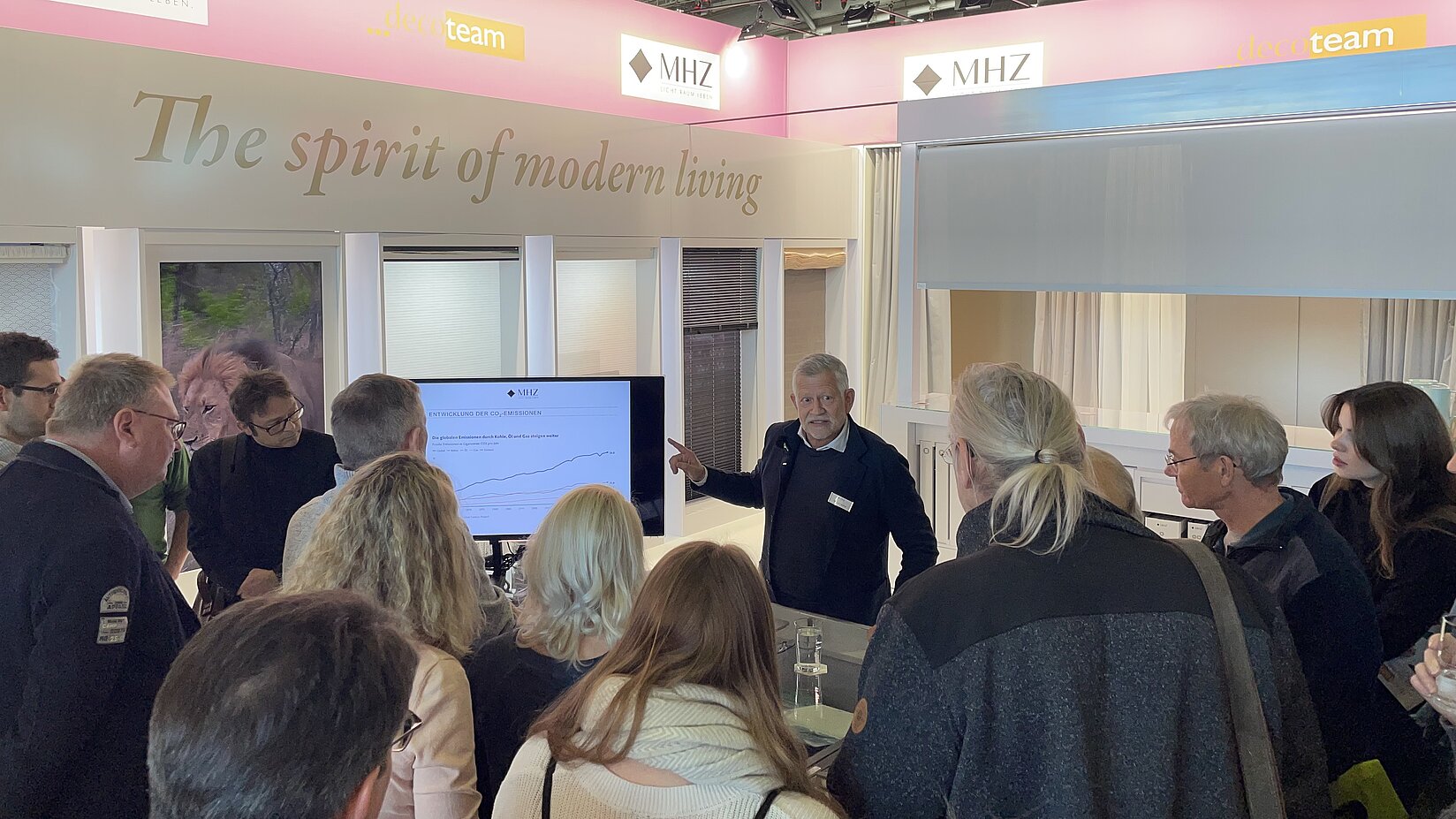 MHZ-Mitarbeiter erklärt Messebesuchern moderne Lösungen für Sicht- und Sonnenschutz am Messestand mit Produktmustern und Bildschirmpräsentation.
