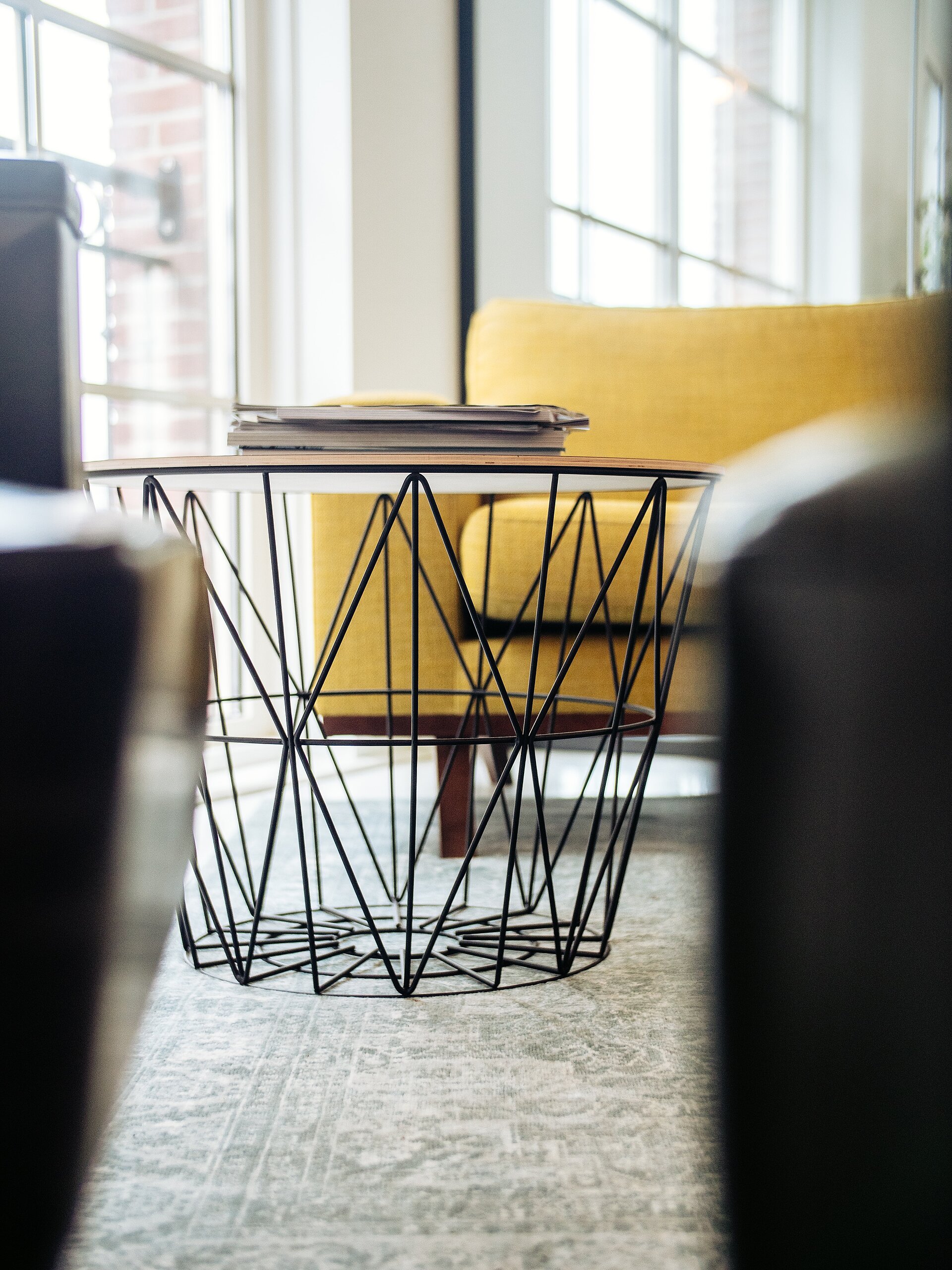 Moderner Wohnraum mit gelbem Sessel und rundem Couchtisch mit schwarzem Drahtgestell und Glasplatte vor grossen Fenstern.