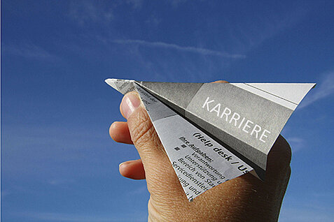 Hand hält Papierflieger mit dem Aufdruck „Karriere“ vor blauem Himmel