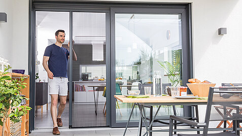 Mann öffnet Insektenschutz-Plissee an einer Schiebetür und geht auf die Terrasse.