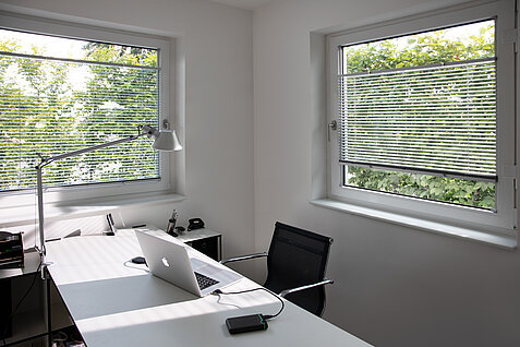 Homeoffice mit zwei Fenstern und silberfarbenen Jalousien. Lamellen regulieren das Licht für blendfreies Arbeiten.