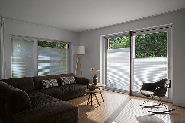 Wohnraum mit dunklem Sofa, weissen Plissees an den Fenstern und Schaukelstuhl in heller Atmosphäre.