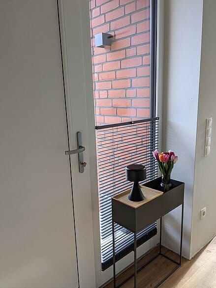 Eingangstür von Innen mit bodentiefen Fenster daneben, das halb mit einer frei verschiebbaren Jalousie verdeckt ist.