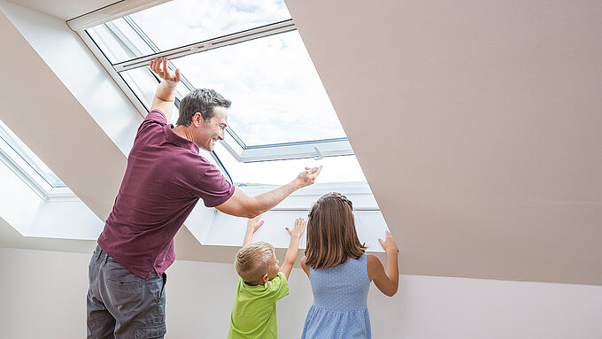 Vater mit zwei Kindern öffnet Dachfenster mit Insektenschutzrollo im Dachgeschoss.