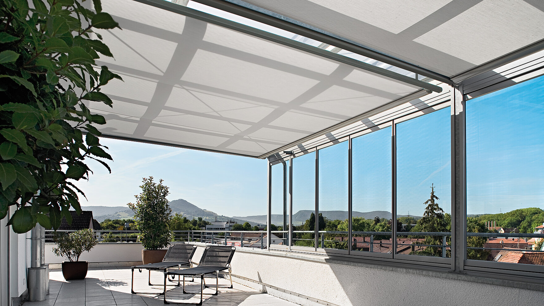 Innenliegende Markisenanlage unter Glasdach einer Dachterrasse mit Aussicht ins Grüne.