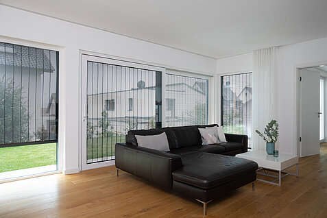 Helles Wohnzimmer mit grossem Fenster, schwarzem Ledersofa und MHZ Metallbehang sonro als Sicht- und Sonnenschutz.