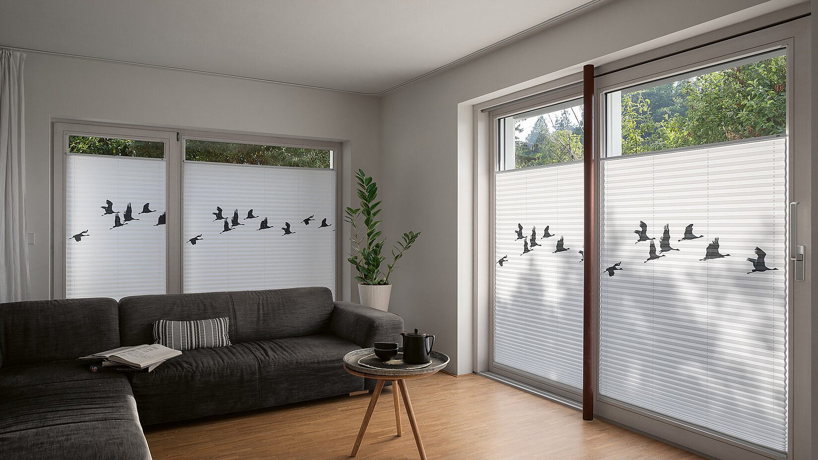Wohnzimmer mit weissen Plissees, die mit einem schwarzen Vogelschwarm-Motiv bedruckt sind.