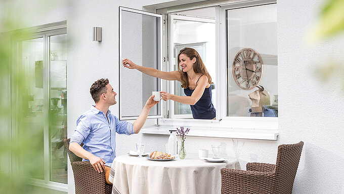 Frau reicht Mann eine Tasse durch ein geöffnetes Pendelfenster mit Insektenschutz.