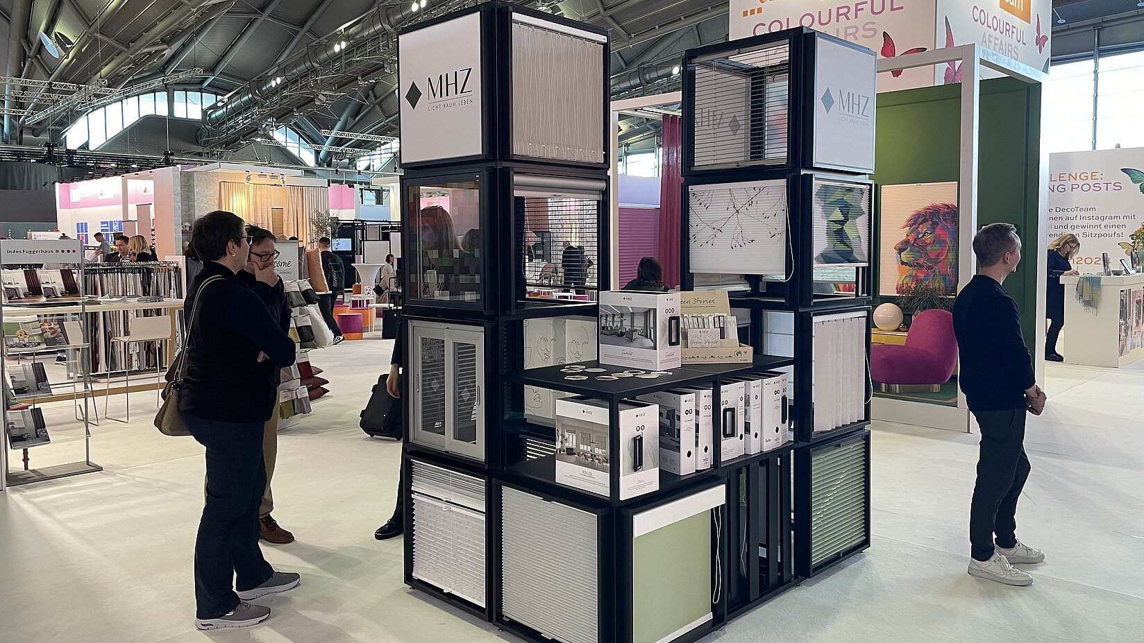 Präsentationsmodul auf dem MHZ-Messestand mit Mustern für Sicht- und Sonnenschutzprodukte, umgeben von Besuchern auf der Messe.