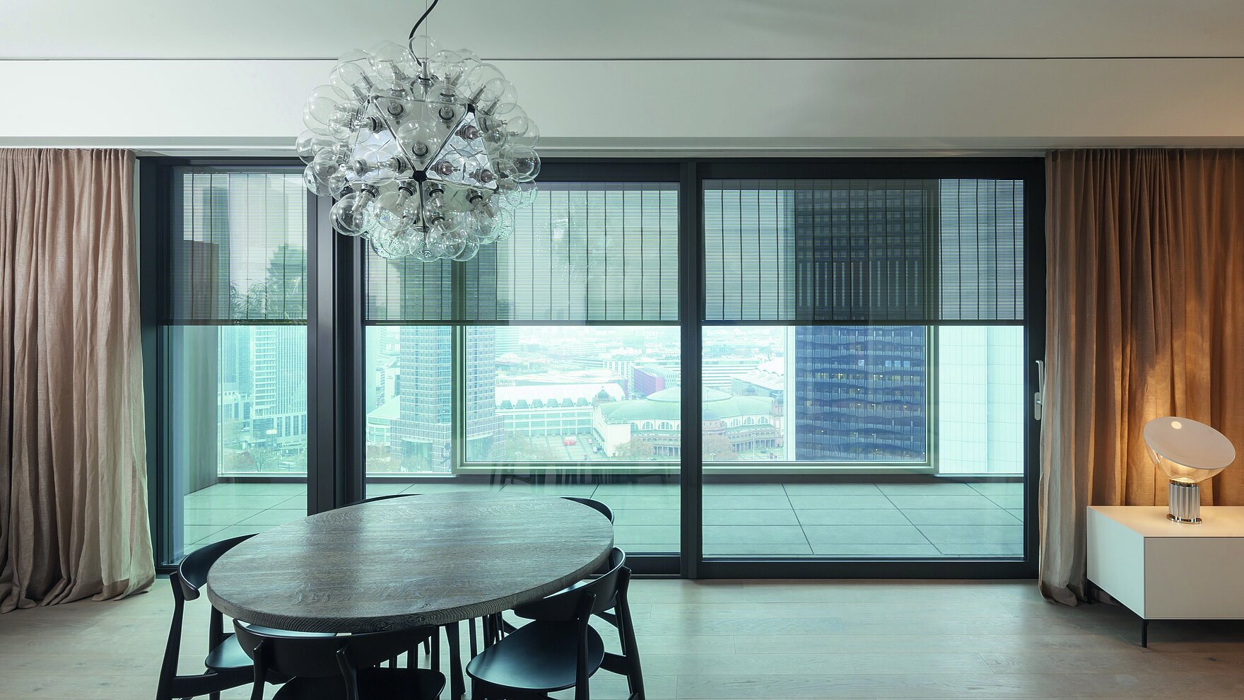 Blick aus modernem Wohnraum mit Esstisch auf grosse Fenster mit aussenliegendem Metallbehang Senro und Stadtblick im Hintergrund.
