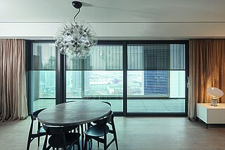 Blick aus modernem Wohnraum mit Esstisch auf grosse Fenster mit aussenliegendem Metallbehang Senro und Stadtblick im Hintergrund.